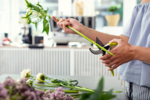 Florist using pruning shears to cut green flower stems on a work table for custom floral arrangements.
